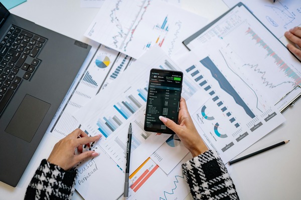Hands holding a smartphone with cryptocurrency trading data next to printed analytics charts
