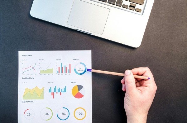Hand pointing a pencil at printed cryptocurrency analytics charts next to a laptop